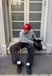 Cappello bordeaux con testo giallo, maglione grigio, pantaloni in velluto a coste neri, calze bianche e scarpe nere. Seduto su gradini di pietra accanto a una porta di vetro.