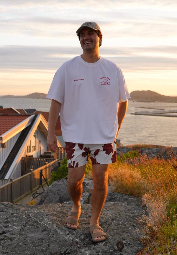 Giovane uomo con cappellino, maglietta bianca e pantaloncini floreali che sorride mentre sta in piedi su terreno roccioso vicino all'acqua al tramonto.