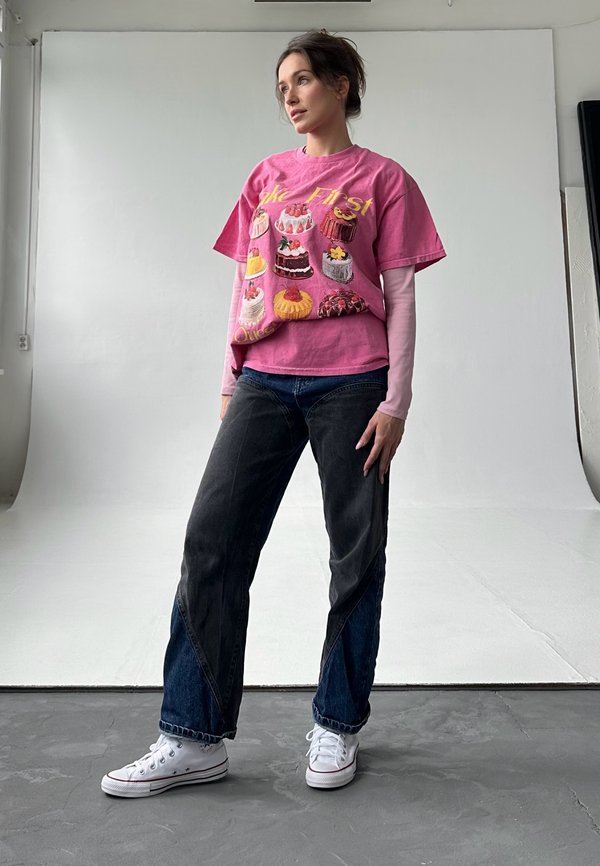 Pink graphic T-shirt med farverige kager, lagd oven på en langærmet bluse. Mørk denim jeans med kontrasterende trekantede paneler, hvide sneakers.