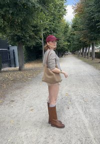 Bottes en cuir marron montant jusqu'au genou, pull beige, et sac à main beige. Le mannequin se tient sur un chemin en gravier bordé d'arbres sous un ciel nuageux.