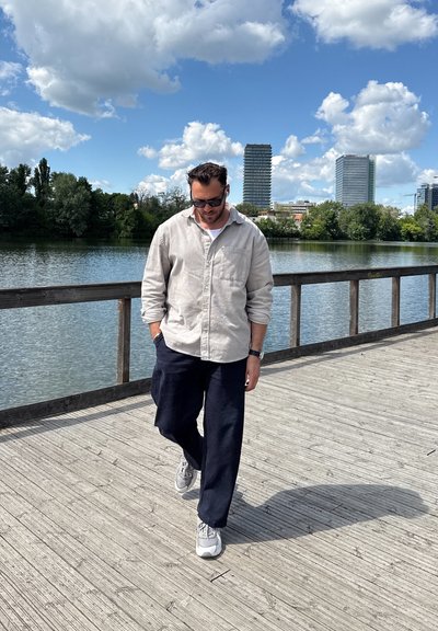 Camisa de botones gris claro, pantalones azul oscuro, zapatillas gris. De pie en un muelle de madera junto al agua, con edificios y árboles al fondo.