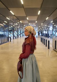 Femme portant un foulard, un chemisier rouge et une longue jupe bleu clair, tenant un sac, debout dans un hall moderne et spacieux avec un éclairage vif au plafond.