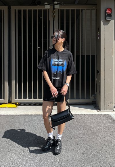 Camiseta gráfica negra con diseño azul, combinada con pantalones cortos negros y sneakers negros gruesos. Los accesorios incluyen un bolso negro de hombro y gafas de sol.