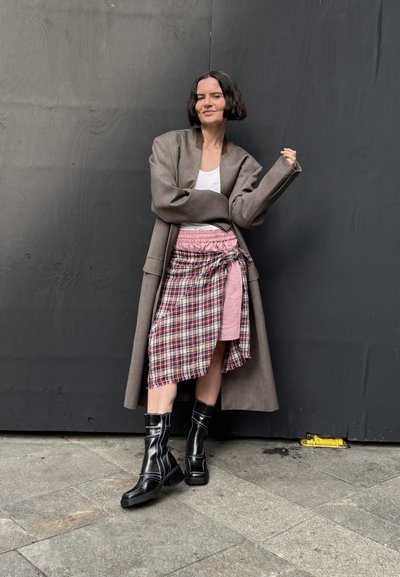Abrigo marrón oversized, camiseta blanca sin mangas, falda rosa con superposición de cuadros y botas negras hasta la rodilla con suela gruesa, de pie sobre una superficie de piedra.