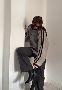Sweater oversized cinza e creme com riscas vermelhas e pretas, calças largas cinzas e botas de tornozelo pretas. Um lenço estampado vermelho e preto está pendurado.