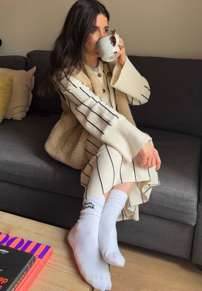 Suéter de punto crema con rayas negras, chaleco de forro polar beige y calcetines blancos. Sentado en un sofá gris con un libro y una taza.