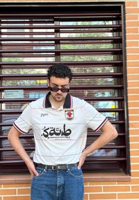 Tricou alb cu mânecă scurtă, guler negru, accente roșii și verzi, și text în arabă. Purtați cu blugi albaștri și curea neagră. Ochelari de soare negri.