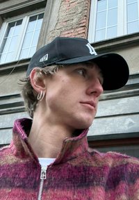 Veste à col et fermeture éclair en tissu à carreaux roses et violets ; portant une casquette noire avec un logo blanc ; boucles d'oreilles argentées en forme d'anneau ; profil latéral partiel.