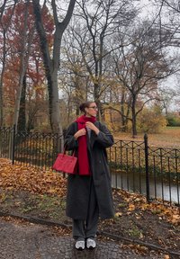 Siv dolg plašč, rdeč šal, rdeča torbica, sive hlače in bele superge. Jesenske liste in drevesa v parku v ozadju z ograjo.