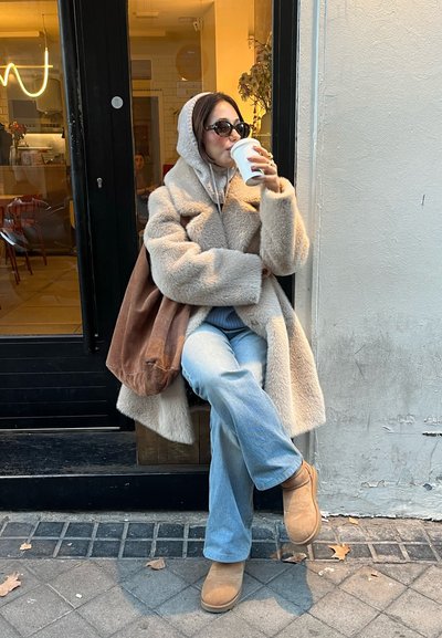 Abrigo de pelo sintético beige con cuello sobredimensionado, sudadera gris, jeans acampanados azul claro, botas de ante color beige y bolso marrón. Sosteniendo una taza blanca.