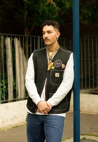 Zwarte denimvest met zijzakken, witte blouse met lange mouwen, blauwe jeans en een gele bandana. Voorzien van insignes en speldjes op het vest.