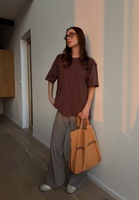 Yli suuri ruskea T-paita, jossa on logo, löysät harmaat housut ja beige liukukengät; pitää käsissään oranssia kantokassia, jossa on mustaa tekstiä.