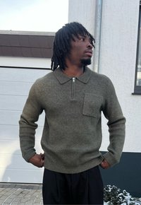 Homme avec des dreadlocks portant un pull zippé vert olive et un pantalon noir, debout en extérieur près d'un bâtiment blanc et de plantes couvertes de neige.
