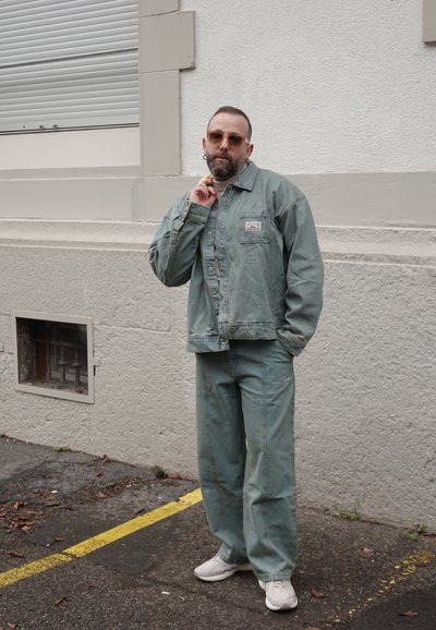 Hombre con barba y gafas de sol, vestido con una chaqueta y pantalones de mezclilla verdes, posando de manera casual contra una pared blanca con textura al aire libre.