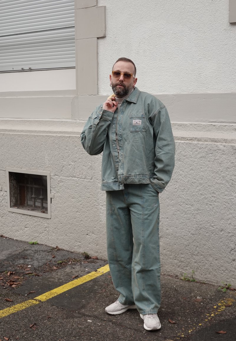 Hombre con barba y gafas de sol, vestido con una chaqueta y pantalones de mezclilla verdes, posando de manera casual contra una pared blanca con textura al aire libre.