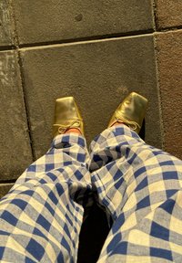 Χρυσά, τετράγωνα παπούτσια με πλεκτή κορδόνι, σε συνδυασμό με μπλε και άσπρα καρό παντελόνια, τοποθετημένα πάνω σε μια υφή πέτρας.