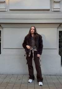 Femme debout contre un mur urbain beige, portant une veste en fourrure marron foncé, une chemise à carreaux nouée à la taille, un pantalon foncé et des baskets blanches.