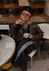 Barna bőrkabátot, fekete beretét és napszemüveget viselő személy ül egy kávézó asztalánál, kávéscsészét tart a kezében és mosolyog.