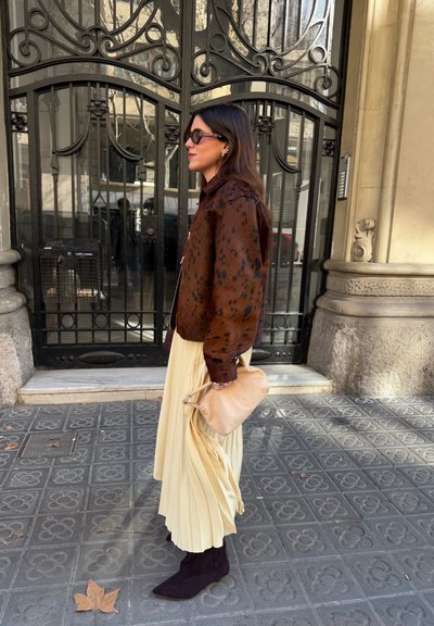 Mujer con chaqueta marrón con manchas, falda plisada crema, botas negras y gafas de sol sostiene un bolso beige mientras está de pie en una acera con patrón junto a una reja negra ornamentada.
