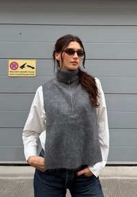 Šedá vesta vyrobená z měkké, texturované tkaniny s vysokým límcem a polovičním zipem. Spárována s bílou pruhovanou košilí a modrými džíny.