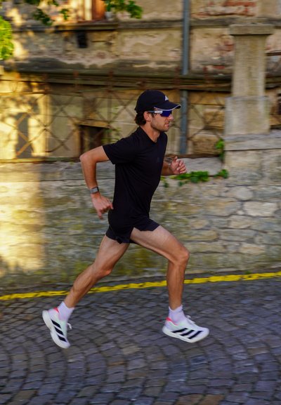Joven vestido con ropa deportiva negra, gorra y gafas de sol corriendo por una calle adoquinada junto a un muro de piedra a la luz del día.