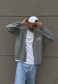 Gaiši pelēks kardigans ar lielām pogām un adītu tekstūru, uz krūtīm ar kontrastējošu baltu burtu, valkāts pār baltu T-kreklu un zilām džinsām.