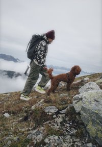 Eine Person in einer gemusterten Daunenjacke, olivfarbenen Cargo-Hosen und leuchtenden Sneakers führt einen lockigen braunen Hund auf steinigem Terrain unter einem bewölkten Himmel.