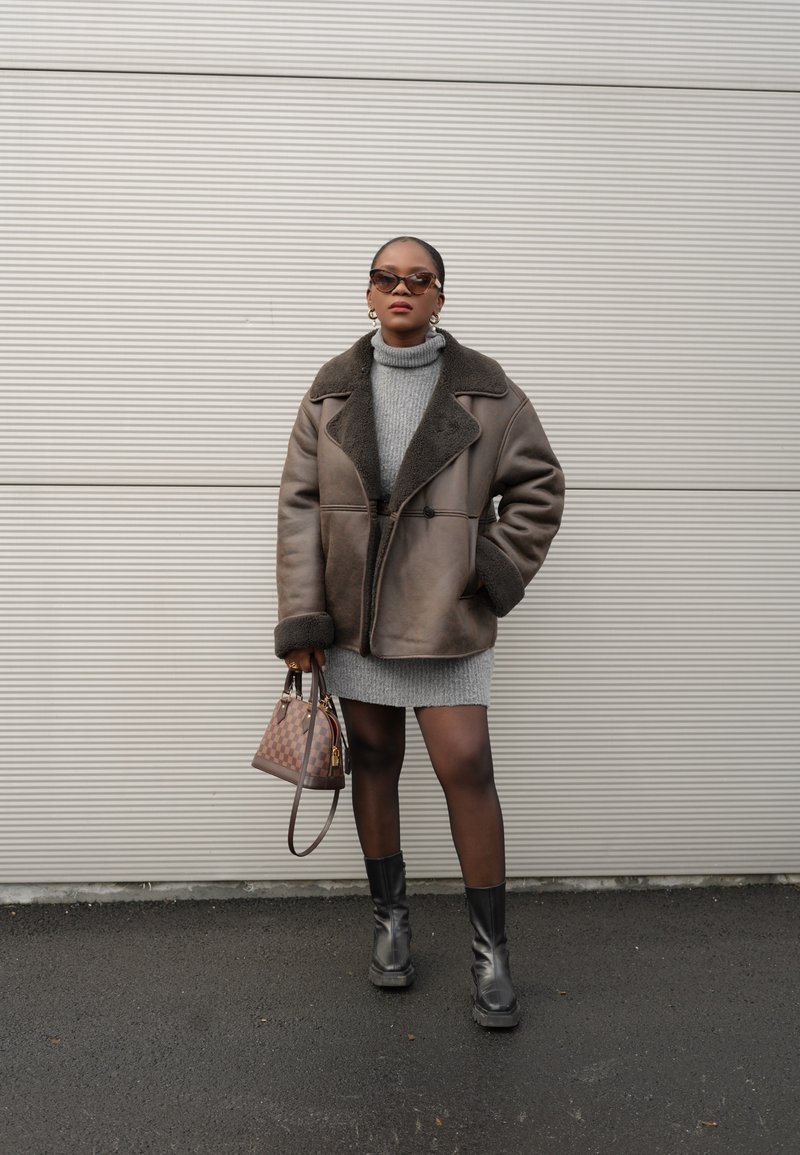 Καφέ γούνινο παλτό με μαύρες λεπτομέρειες, γκρι πλεκτό φόρεμα, μαύρες μπότες ως το γόνατο και καφέ καρό τσάντα. Στερεό φόντο.