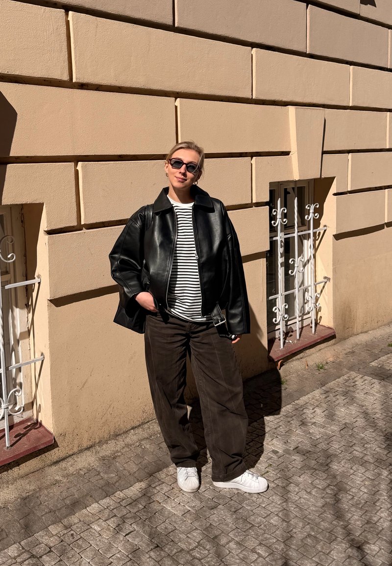 Personne portant une veste en cuir noire, une chemise rayée, un pantalon foncé et des baskets blanches, debout dans une rue pavée à côté d’un mur beige avec des fenêtres grillagées.