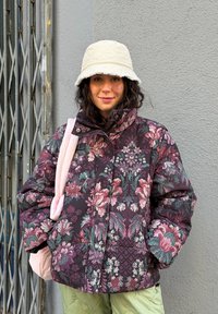 Dunjakke med blomstertrykk i mørk lilla og grønt, med høy krage og quiltet tekstur. Kombinert med en lys rosa veske.