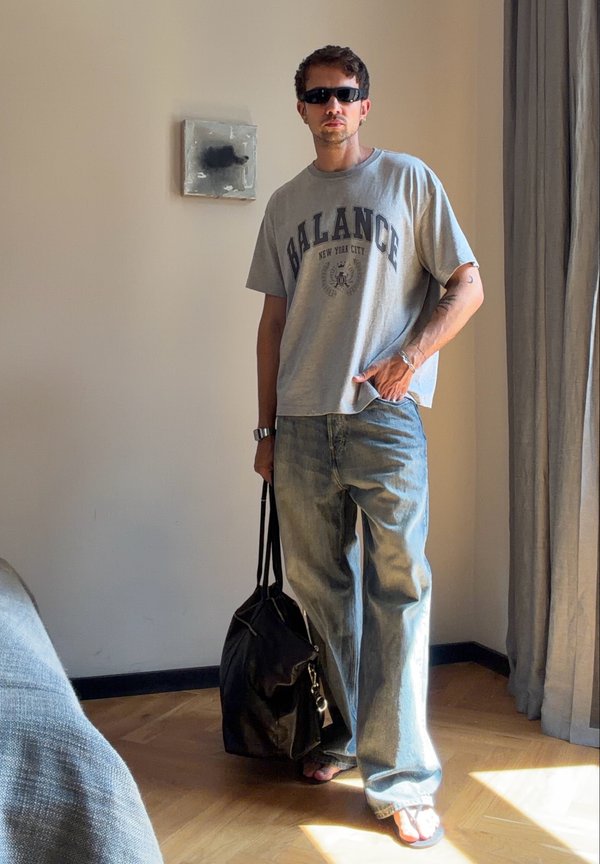 Man wearing sunglasses, loose gray balance t-shirt, baggy jeans, and sandals holding a black bag standing in a sunlit room.