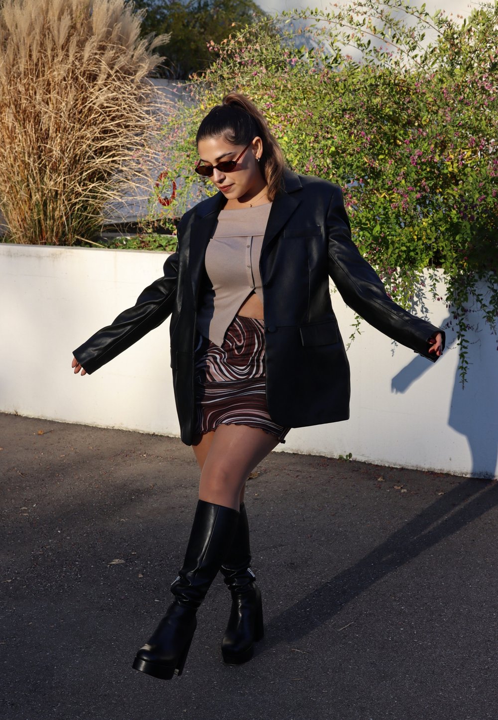 Svart läder oversized blazer, beige kort topp, mönstrad mini kjol med swirlar, och knähöga svarta klackboots, mot en grönskande bakgrund.
