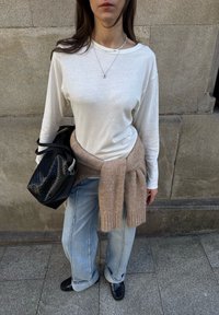 Jasnoszara koszula z długim rękawem, beżowy sweter związany wokół talii, luźne jasnoniebieskie dżinsy, czarna teksturowana torba, czarne buty. Neutralne tło.