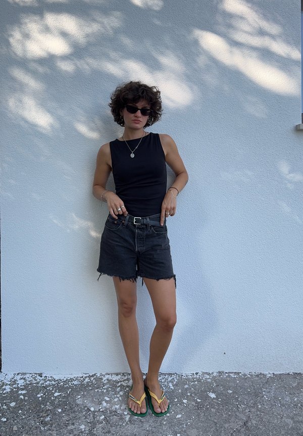 Persona con cabello rizado, lleva una camiseta negra sin mangas, pantalones cortos de mezclilla negros, gafas de sol y chanclas, está de pie contra una pared blanca.