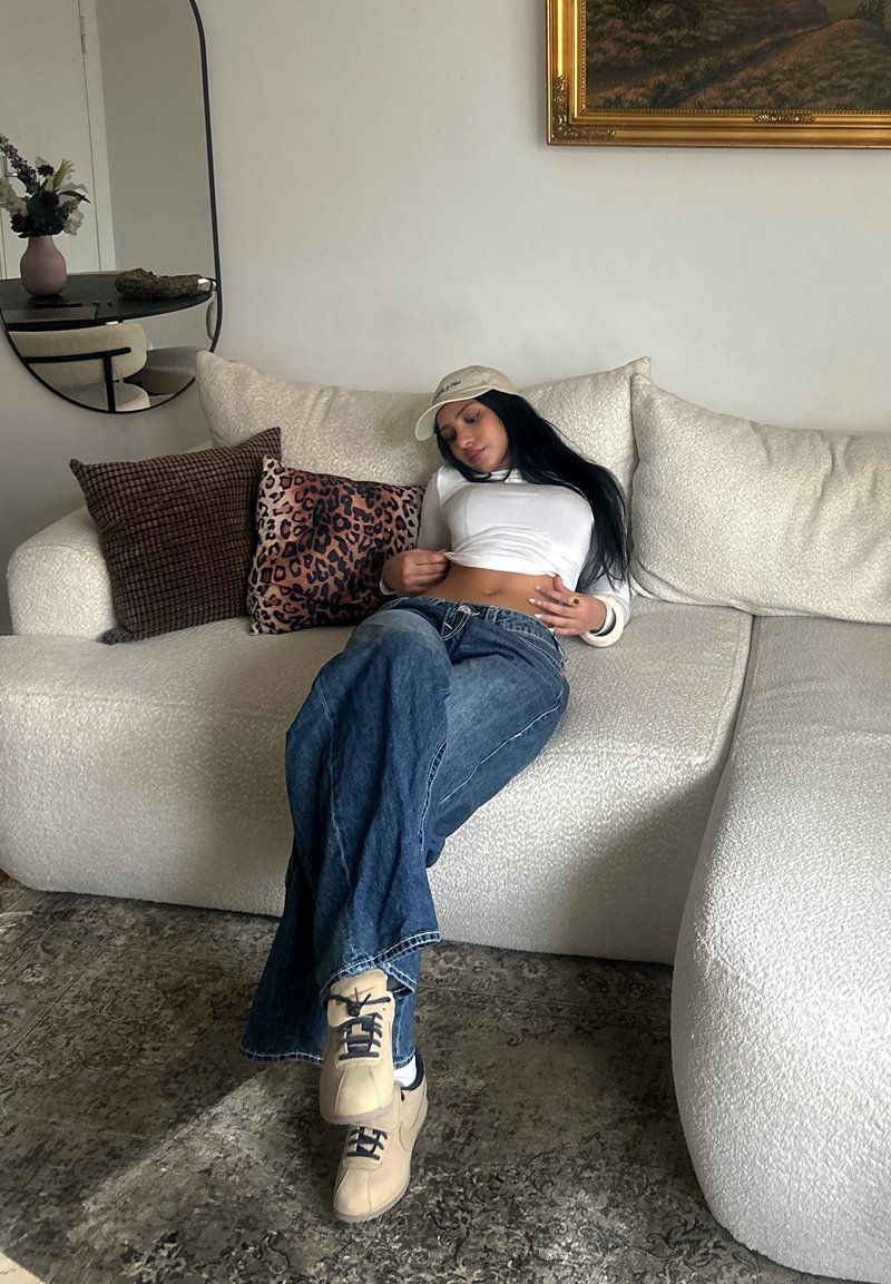 Femme portant une casquette, un crop top blanc et un jean, allongée sur un canapé blanc avec des coussins à motifs dans un salon moderne.