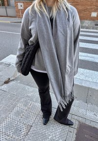Personne debout sur un trottoir en ville au passage piéton, portant une écharpe grise, un pull gris, un pantalon noir, des chaussures noires, et tenant un sac matelassé noir.