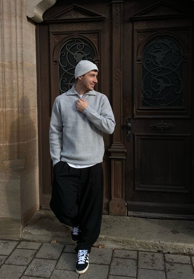 Suéter gris de canalé con un cuello alto, combinado con pantalones negros holgados y zapatillas deportivas. El fondo presenta puertas de madera ornamentadas con detalles decorativos.