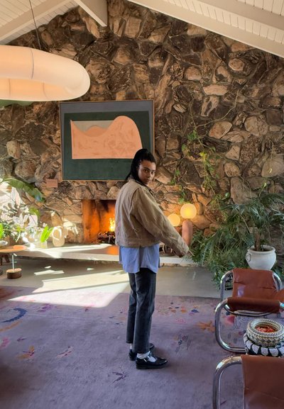 Chaqueta marrón de gran tamaño, camisa azul clara, jeans oscuros y zapatos negros. Fondo de pared de piedra, chimenea cálida y plantas en un interior moderno.