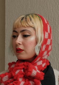 Roter und rosa kariert gestrickter Schal mit rüschiger Textur, als Kapuze getragen, kombiniert mit einem schwarzen Kleidungsstück. Kurzes, blondes Haar mit gefärbten Spitzen.