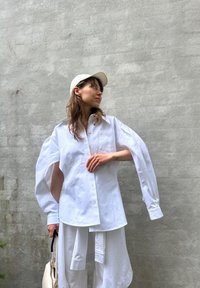Camisa branca de botão com mangas oversized, bolso frontal e textura suave, combinada com calças brancas e um boné de cor clara.
