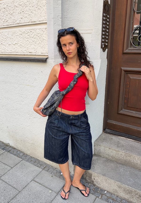 Jeune femme en débardeur rouge et short en jean foncé, debout sur un trottoir près d'une entrée de bâtiment avec un sac à épaule gris à motifs.
