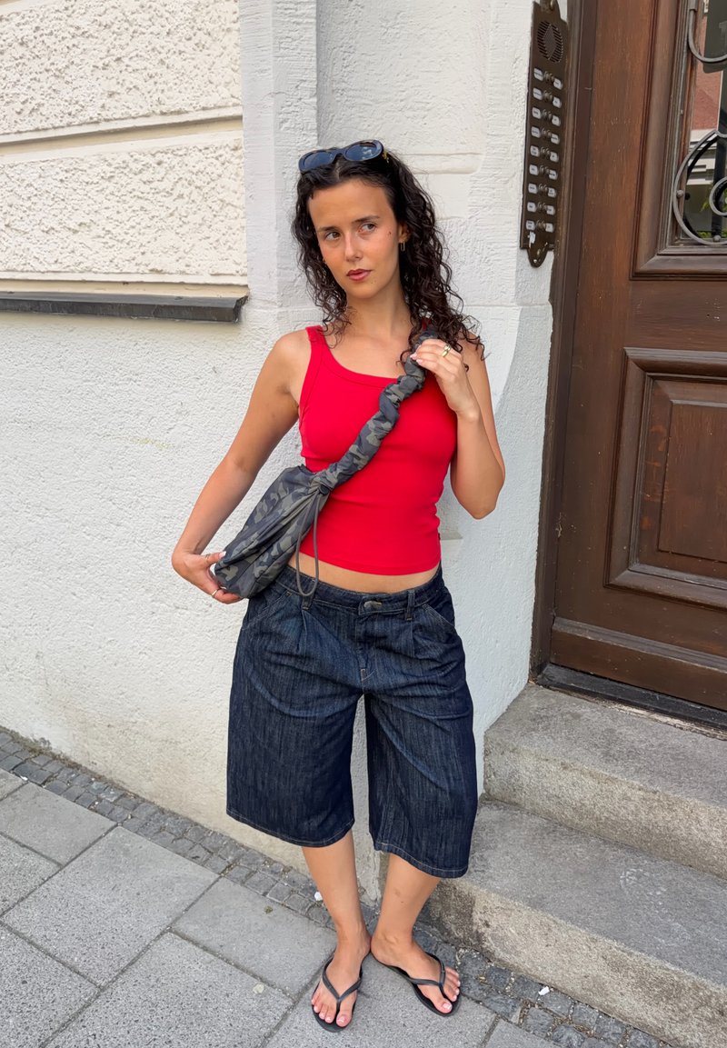 Jeune femme en débardeur rouge et short en jean foncé, debout sur un trottoir près d'une entrée de bâtiment avec un sac à épaule gris à motifs.