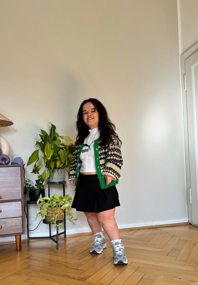 Cárdigan de punto en patrones crema y negro con acentos verdes, camiseta cropped blanca, falda negra y zapatillas grises con calcetines blancos. Entorno interior.