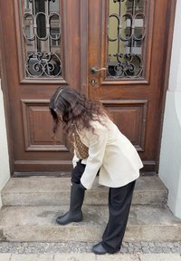 Black rubber boot with a rounded toe, worn with dark trousers and a light blazer, positioned on a stone step in front of a wooden door.