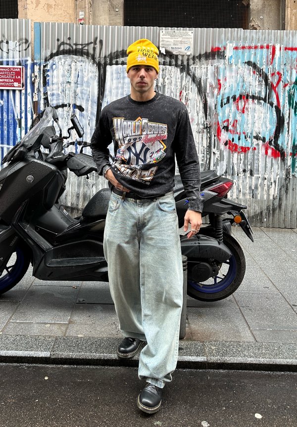 Mann mit gelber Mütze und schwarzem NY Yankees Sweatshirt steht vor einem schwarzen Roller vor einem mit Graffiti bedeckten Metallzaun.