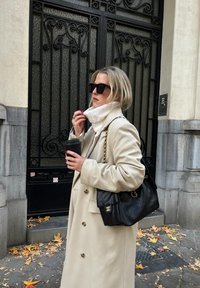 Femme portant un manteau beige et un col roulé blanc, tenant une tasse de café et un sac à main noir matelassé, debout sur un trottoir urbain couvert de feuilles près d'une porte ornée.