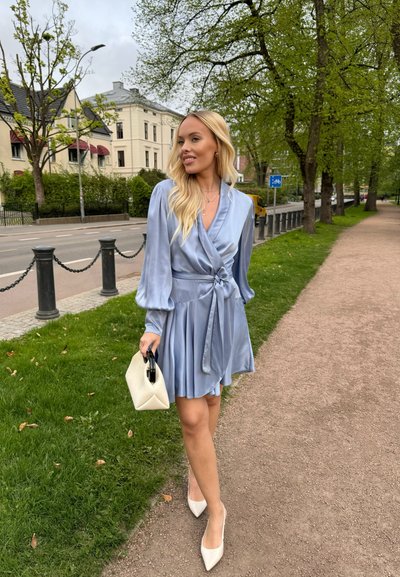 Vestido de satén azul claro con mangas largas abullonadas, diseño envolvente y falda plisada. Combinado con un bolso geométrico crema y tacones blancos.