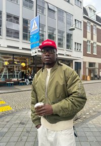 Homme avec une casquette rouge et une veste olive tenant une tasse de café dans une rue urbaine avec un café et un panneau de passage pour piétons en arrière-plan.