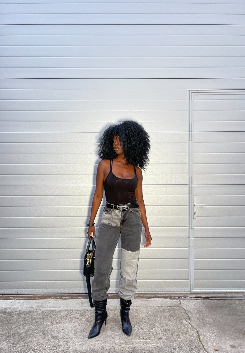 Schwarzer Bodysuit, graue und hellgraue Patchwork-Jeans, schwarze spitze Stiefel und eine schwarze Handtasche, vor einem hellgrauen strukturierten Hintergrund.