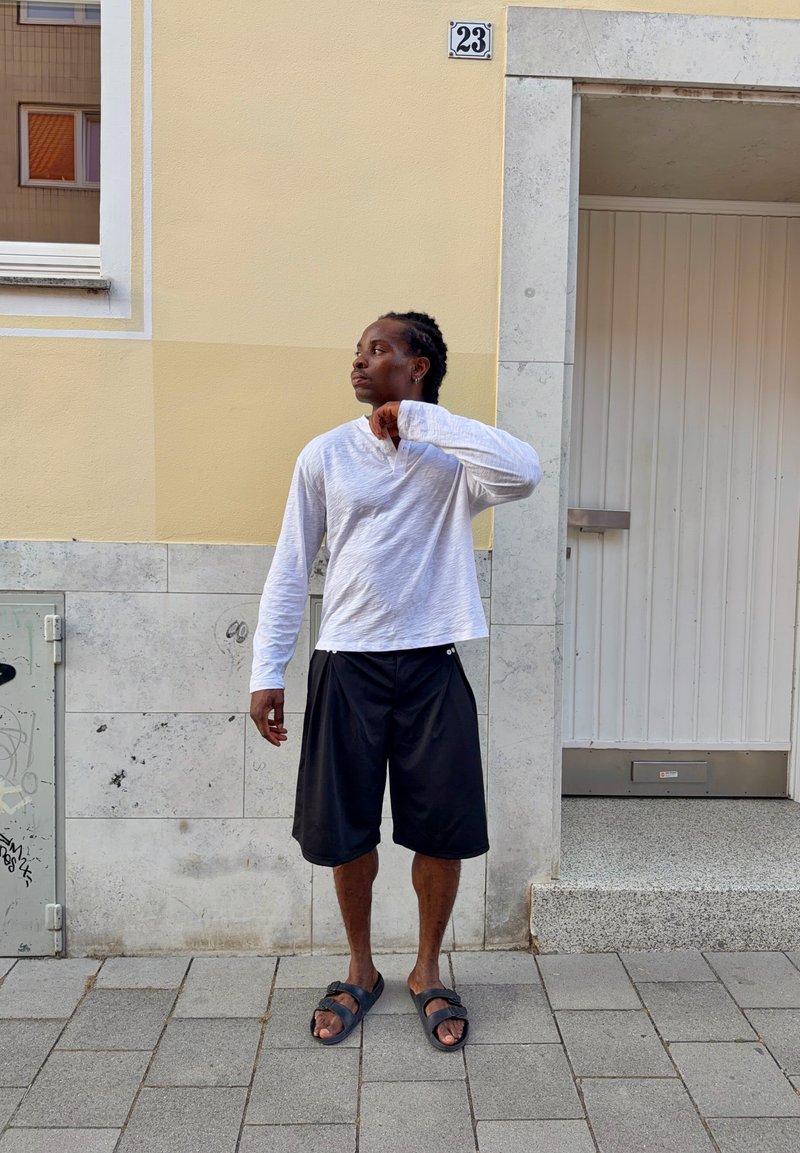 Homme portant une chemise blanche à manches longues, un short noir et des sandales, debout sur le trottoir, regardant sur le côté près d’un bâtiment avec une porte et une fenêtre.